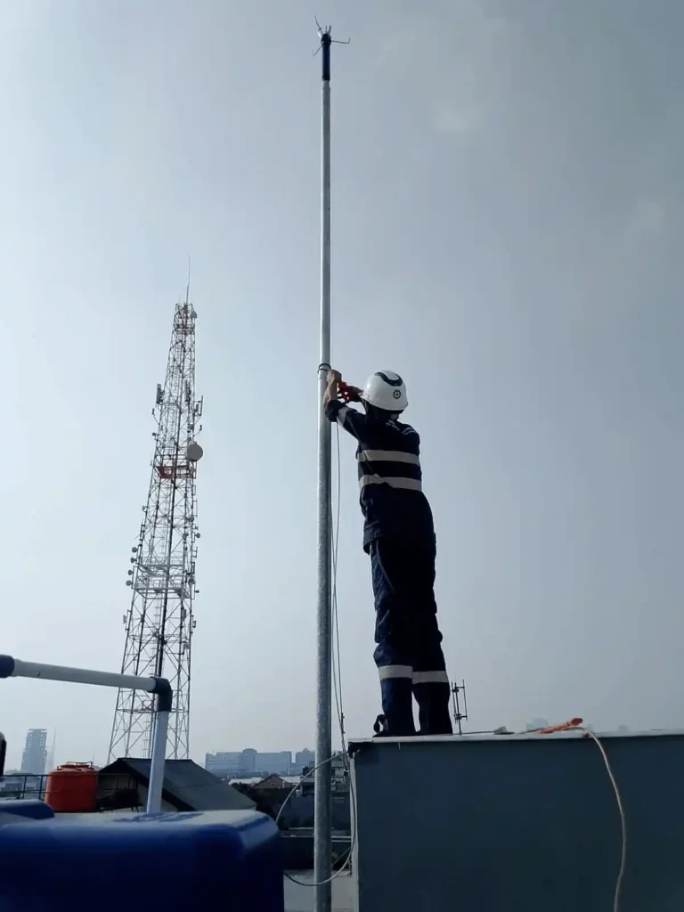 “Teknisi memasang tiang penangkal petir di atap gedung dengan menara telekomunikasi di latar belakang.”